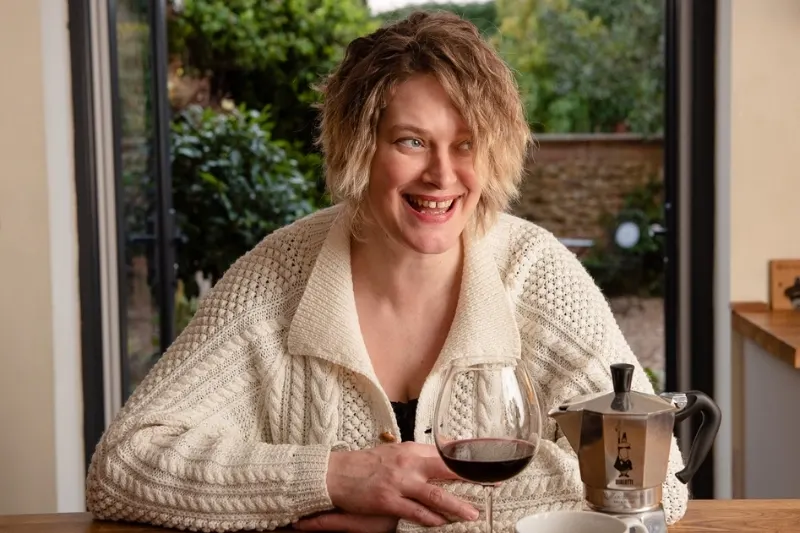 Woman wearing a hand knitted cream aran knit cardigan with a glass of wine and coffee pot beside her.
