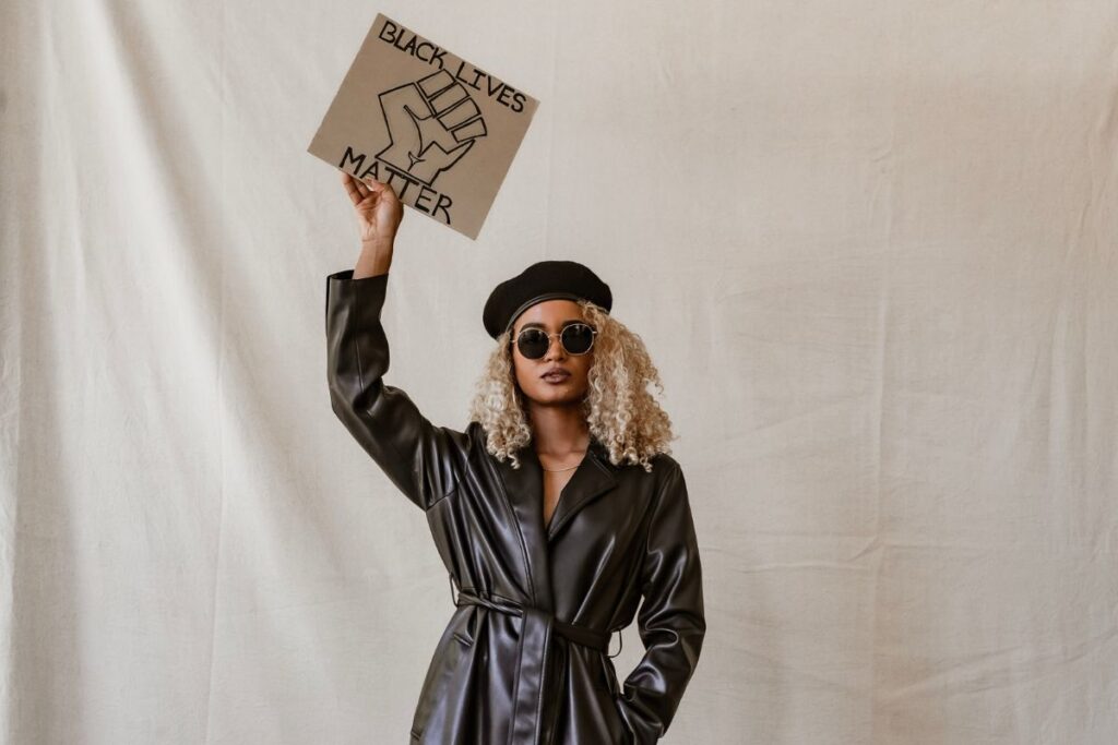 Black woman wearing a black leather style trench coat holding a card placard with 'Black Lives Matter' written on!